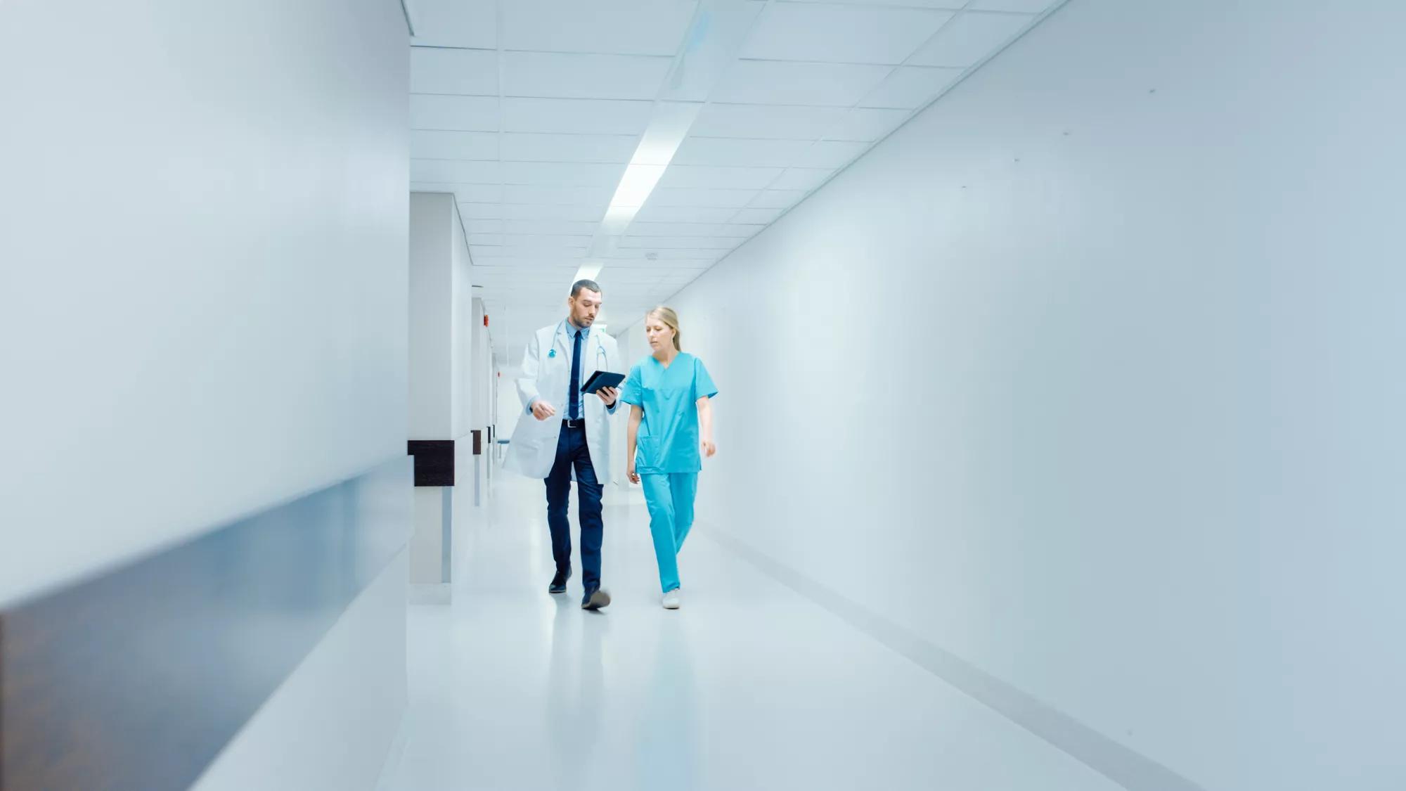 Surgeon and Female Doctor Walk Through Hospital Hallway in a Hurry while Using Digital Tablet and Talking about Patient's Health. Modern Bright Hospital with Professional Staff.