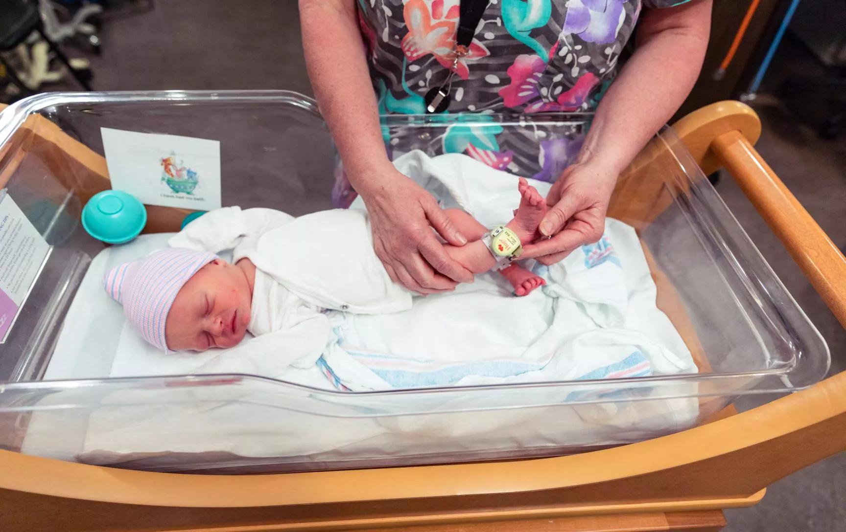 Caregiver showing Hugs Infant Protection Wi-Fi badge on a newborn's ankle in a clear bassinet.