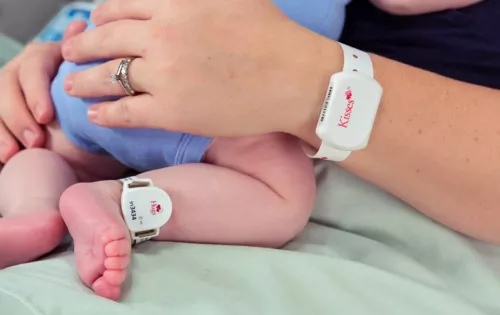 Mom holding her newborn baby with the Hugs Infant badge on baby's ankle, while wearing the Kisses Mother band on her wrist.