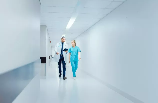 Surgeon and Female Doctor Walk Through Hospital Hallway in a Hurry while Using Digital Tablet and Talking about Patient's Health. Modern Bright Hospital with Professional Staff.