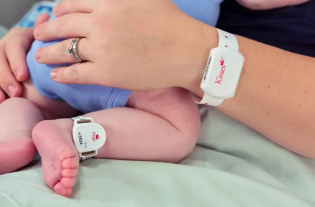 Mom holding her newborn baby with the Hugs Infant badge on baby's ankle, while wearing the Kisses Mother band on her wrist.