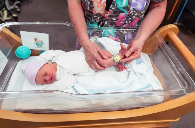 Caregiver showing Hugs Infant Protection Wi-Fi badge on a newborn's ankle in a clear bassinet.