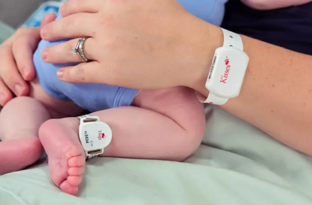 Mom holding her newborn baby with the Hugs Infant badge on baby's ankle, while wearing the Kisses Mother band on her wrist.
