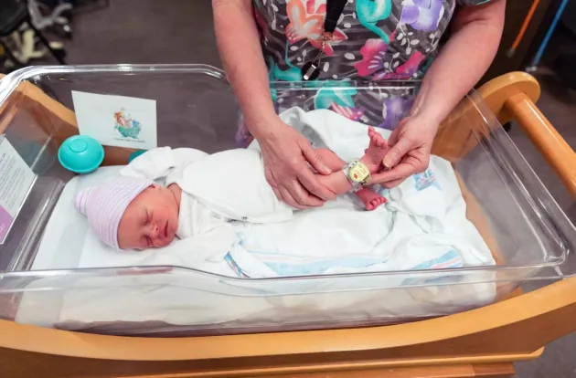 Caregiver showing Hugs Infant Protection Wi-Fi badge on a newborn's ankle in a clear bassinet.